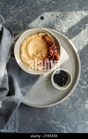 Vista dall'alto dell'hummus ricoperto di jamon a fette, servito in una ciotola di ceramica su un tavolo di marmo. L'impostazione include una piccola ciotola di semi di sesamo nero. Foto Stock