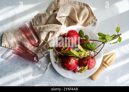 Vista dall'alto di una serie di melograni maturi disposti su un piatto bianco, accompagnati da foglie verdi decorative e motivi d'ombra. Bicchieri rosa e una biancheria Foto Stock