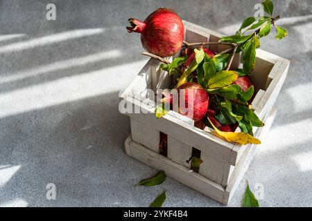 Un'immagine laterale che mostra melograni maturi con foglie verdi vibranti in una cassa di legno bianca, gettata nelle delicate ombre della luce del sole. Foto Stock