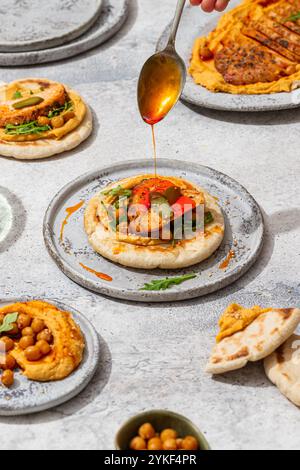 La vista dall'alto mostra un piatto colorato di pollo arrosto servito su hummus guarnito con verdure, condite con olio, circondato da naan e ceci freschi Foto Stock