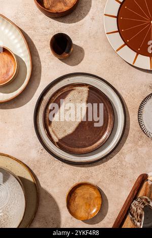 Vista dall'alto di una splendida collezione di piatti, ciotole e bicchieri in ceramica artigianali, elegantemente stenditi su una superficie del tavolo illuminata dal sole e strutturata. Il variegato Foto Stock