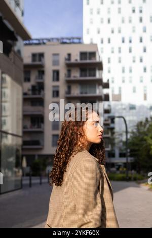 Una giovane donna d'affari brasiliana in un blazer elegante cammina con fiducia in un ambiente urbano moderno, riflettendo professionalità e determinazione Foto Stock