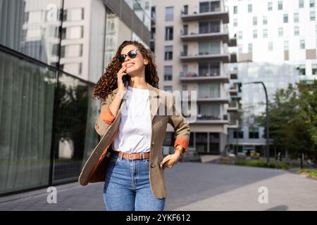 Un'elegante donna d'affari brasiliana cammina con sicurezza mentre parla sul suo smartphone fuori dagli edifici moderni è vestita con un blazer chic e una sungla Foto Stock
