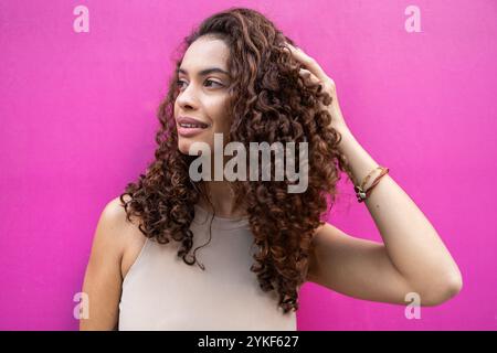 Donna d'affari brasiliana sicura di sé con capelli ricci sorridenti, posta su un vibrante sfondo rosa, trasuda un'aura professionale ma avvicinabile Foto Stock