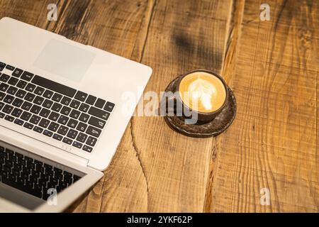 Un computer portatile insieme a una tazza da caffè con latte art, collocato su un rustico tavolo in legno, raffigurante un moderno impianto di lavoro remoto Foto Stock
