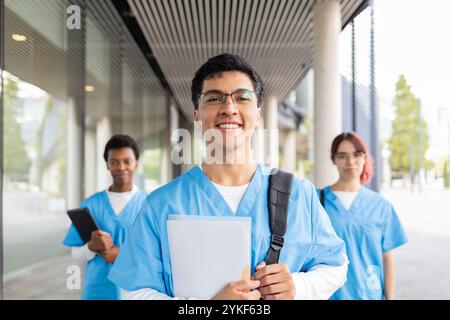 Allegri studenti di allattamento in scrub blu, con libri e borse, camminando con sicurezza fuori dalla loro scuola di medicina, pronti per la loro prossima classe Foto Stock