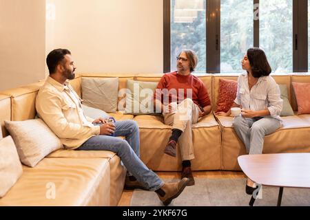 Una riunione rilassata in uno spazio di coworking dove tre persone chiacchierano casualmente su un accogliente divano in pelle con cuscini colorati la luce naturale diffonde la luce Foto Stock