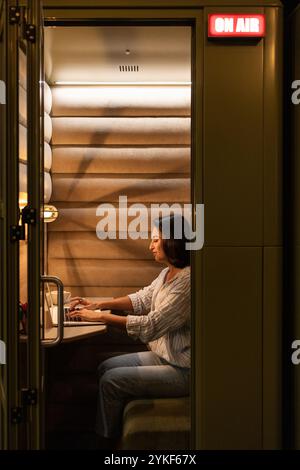 Una donna lavora intensamente sul suo laptop in un accogliente stand insonorizzato in uno spazio di coworking l'illuminazione calda e il segnale aereo creano un'atmosfera di convivialità Foto Stock