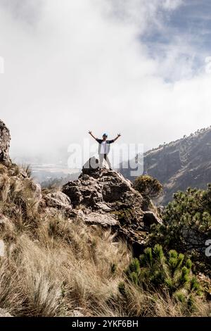 Un escursionista si erge trionfalmente su una cima rocciosa, braccia allungate, circondato da vegetazione lussureggiante e un cielo nuvoloso mozzafiato, a simboleggiare lo spirito di un Foto Stock