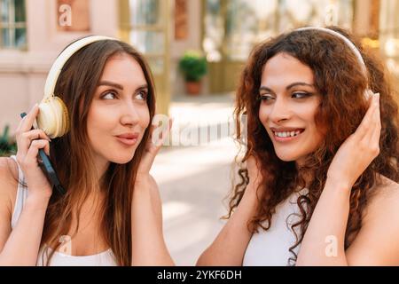 Una donna transessuale e la sua amica vengono raffigurate mentre ascoltano musica all'aperto, condividono sorrisi indossando le cuffie in un ambiente urbano e soleggiato Foto Stock