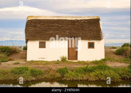 Un caratteristico cottage bianco con tetto in paglia adagiato sullo sfondo pittoresco del Delta dell'Ebro in Spagna, che si riflette in un corpo idrico Foto Stock