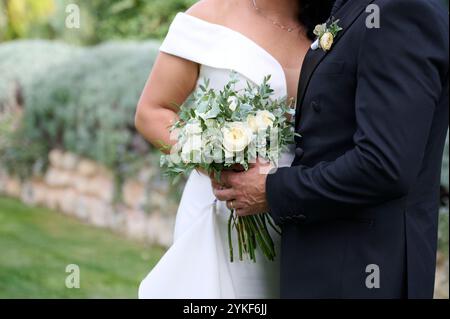 Vista corta e irriconoscibile di una sposa in un abito bianco a spalla e di uno sposo in una tuta nera, con un bouquet di rose bianche e una vegetazione lussureggiante, Foto Stock