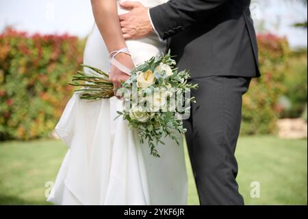 Vista corta di una sposa e di uno sposo che si tengono per mano durante la cerimonia di nozze, concentrandosi sull'elegante bouquet nuziale di fiori bianchi e verdi. Il Foto Stock