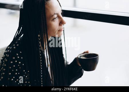 Un'elegante e premurosa donna caucasica con trecce viene catturata bevendo caffè in un caffè. Sembra serena, guardando lontano dalla telecamera mentre assapora Foto Stock