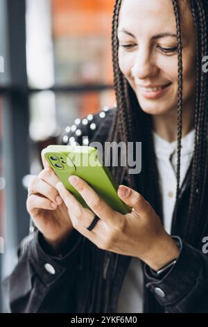 Un'elegante donna caucasica con trecce sorride mentre usa il suo smartphone in un bar moderno, mostrando un momento di uso informale della tecnologia nella vita di tutti i giorni Foto Stock