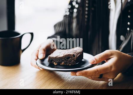 Vista ravvicinata delle mani di una persona anonima che serve una fetta di brownie su un piatto nero, con una tazza di caffè nelle vicinanze, posizionata su un tavolo di legno. Concentrati su Foto Stock