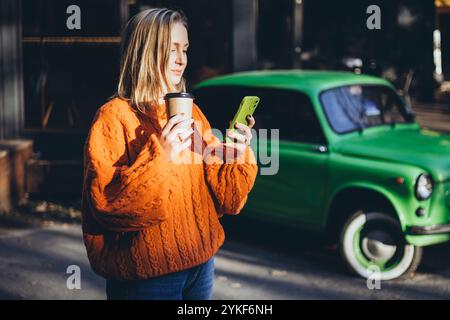 Una donna con un comodo maglione in maglia arancione regala una tazza di caffè e uno smartphone, mentre mentre si prende il sole sullo sfondo si siede Un'auto verde d'epoca, aggiungendo il carattere Foto Stock