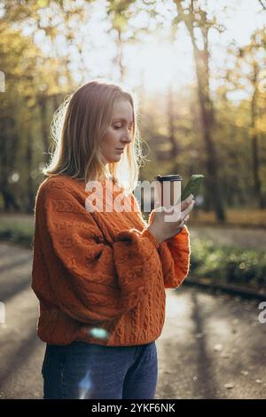 Una donna che indossa un caldo maglione arancione lavorato a maglia si trova nella luce soffusa del sole che filtra tra gli alberi autunnali e tiene in mano una tazza di caffè e uno smartphon Foto Stock