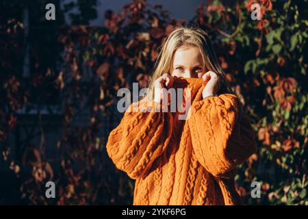 Una donna nasconde giocosamente il viso in un caldo e accogliente maglione arancione lavorato a maglia. La ricca texture del maglione completa le vivaci foglie autunnali nella b Foto Stock