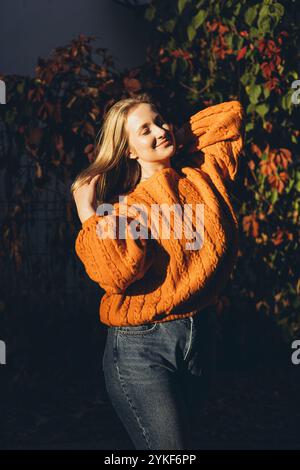 Una donna con un vivace maglione a maglia arancione si crogiola nella calda luce autunnale, circondata da vegetazione lussureggiante e fogliame rosso, creando un ambiente accogliente e sereno Foto Stock