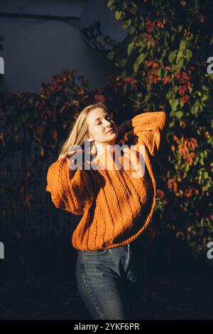 Una donna si crogiola nel calore di una giornata illuminata dal sole, indossando un comodo maglione in maglia arancione sullo sfondo di vivaci foglie autunnali si completa con il suo stile rilassato Foto Stock