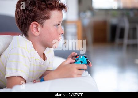 Un ragazzo concentrato si siede comodamente su un divano, giocando intensamente a un videogame utilizzando un controller, guardando lontano dalla telecamera. Foto Stock