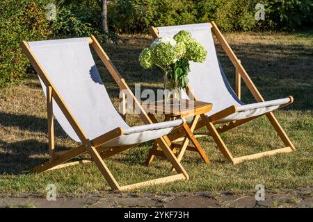 Una tranquilla terrazza con due sedie a sdraio e un piccolo tavolo in legno con un vaso di fiori freschi, che rappresenta un tranquillo salotto e relax Foto Stock