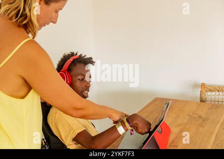 Un giovane ragazzo afroamericano con paralisi cerebrale si impegna in un'attività di apprendimento utilizzando un tablet nella sua casa di vacanza, guidato dalla sua insegnante femminile. Lui io Foto Stock