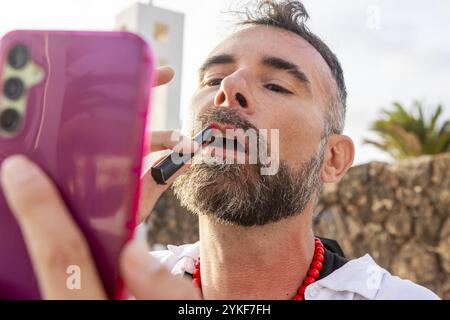 Un gay travestito che applica con sicurezza il rossetto mentre usa il suo telefono come specchio all'aperto, incarnando l'espressione LGBTQIA+ Foto Stock