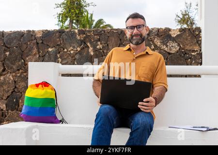 Un uomo gay sicuro di sé con occhiali e una camicia dorata utilizza un computer portatile seduto su una panchina bianca con una colorata borsa color arcobaleno Pride accanto a lui, che incarna LG Foto Stock