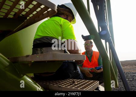 Due lavoratori, che indossano caschi e giubbotti ad alta visibilità, sono visti alla base di una turbina eolica in un parco eolico impegnato in discussioni, dimostrano Foto Stock