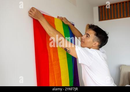 Un giovane trans appende una colorata bandiera d'orgoglio sul suo muro bianco, decorando la sua nuova casa ed esprimendo la sua identità Foto Stock