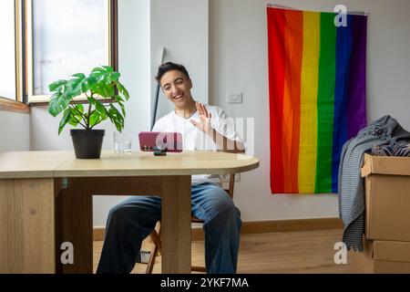 Un uomo trans videochiama utilizzando uno smartphone, sventolando felicemente dalla sua nuova casa adornata da una bandiera d'orgoglio Foto Stock