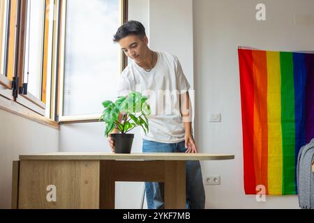 Un uomo trans prepara la sua casa, posizionando una pianta in vaso sul tavolo con una vibrante bandiera Pride sullo sfondo, che rappresenta la sua identità e la LGBTQ Foto Stock