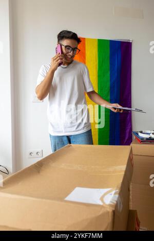 Un uomo trans parla al telefono tenendo in mano un appunti, sorridendo in una stanza decorata con una vibrante bandiera d'orgoglio, circondata da scatole mobili Foto Stock