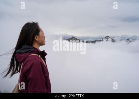 Una giovane donna con una giacca Bordeaux si erge in modo contemplativo su uno sfondo di cime di montagna ricoperte di nuvole nel tranquillo paesaggio di Stoos, Switzer Foto Stock