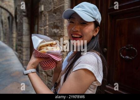 Una donna asiatica assapora il tradizionale cibo di strada italiano mentre esplora le affascinanti strade lastricate di pietra di Volterra, in Toscana, la sua delizia riflette il Foto Stock
