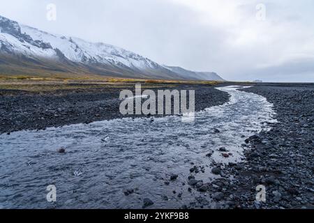 Un tranquillo fiume si snoda attraverso il paesaggio roccioso delle Highlands islandesi le montagne innevate si innalzano maestosamente sullo sfondo, sotto una cl Foto Stock