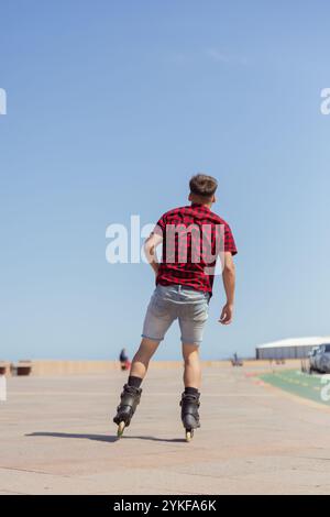 Vista posteriore di un giovane irriconoscibile che cammina a rotelle in una giornata di sole, con una camicia a quadri rossa e pantaloncini in denim. L'immagine mostra l'energia A. Foto Stock