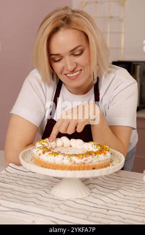 Una donna in grembiule tocca la meringa con una torta adornata da colorate spruzzette ambientate in una cucina accogliente, si gode il momento con un sorriso, sh Foto Stock