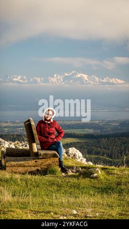 Una donna, guardando la macchina fotografica, siede su una panchina di legno a Dent de Vaulion durante il tramonto, con le Alpi svizzere sullo sfondo sotto un cielo limpido. Foto Stock