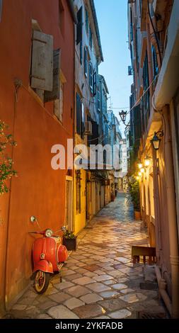 Un pittoresco vicolo di Corfù, Grecia, presenta una moto rossa parcheggiata contro una parete colorata luci calde e strade acciottolate creano un'atmosfera accogliente Foto Stock
