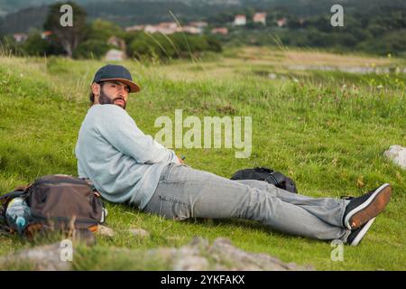 Un uomo con un abbigliamento casual giace rilassato sull'erba, godendo di un paesaggio sereno nelle Asturie, in Spagna, con il suo zaino nelle vicinanze, comunicando la pace di Foto Stock