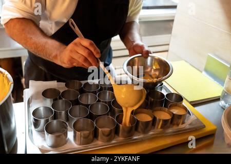 Lo chef professionista versa con cura la pastella in stampi cilindrici per la cottura, in una cucina ben attrezzata del ristorante, che mostra la competenza culinaria e Foto Stock