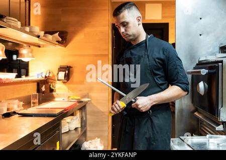 Un giovane chef maschile affilerà un grande coltello da cucina, preparandosi per l'animato servizio di cena in un ristorante contemporaneo ben attrezzato Foto Stock