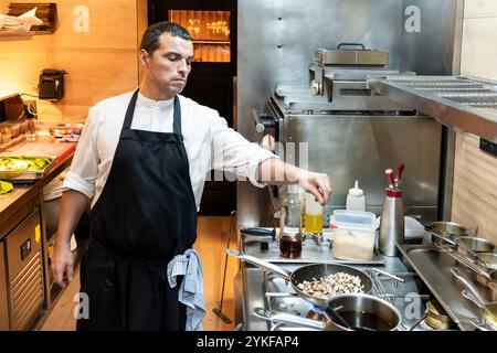 Uno chef maschile dedicato cucina sapientemente in un ristorante contemporaneo, concentrandosi mentre semina un piatto, circondato da attrezzature culinarie professionali Foto Stock