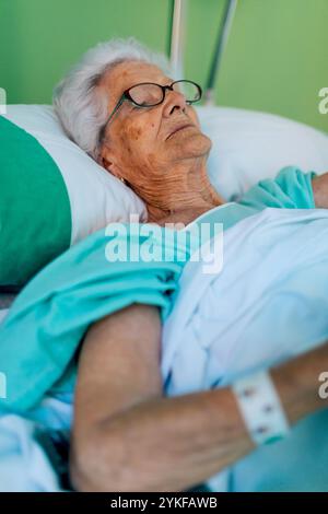 Una donna anziana con gli occhiali giace pacificamente in un letto d'ospedale, ricoperta da un abito da ospedale color tè e lenzuola bianche la sua espressione serena e la calma ambia Foto Stock