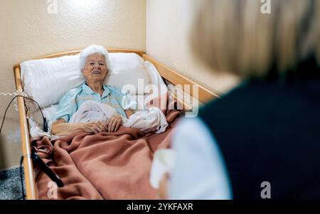 Una donna anziana con i capelli bianchi è sdraiata a letto, sorridente mentre si impegna con un visitatore tonalità calde e coperte accoglienti creano un'atmosfera confortevole Foto Stock