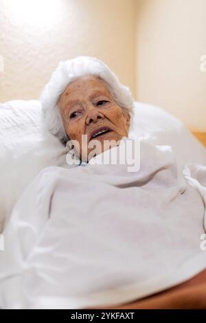 Una donna anziana e serena si trova comodamente a letto, avvolta da una coperta bianca, la sua espressione è calma, incarna pace e tranquillità in una rosa dai toni caldi Foto Stock
