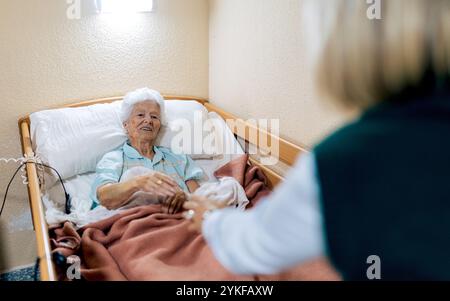 Una donna anziana giace comodamente a letto in una residenza di cura, salutando calorosamente un visitatore la camera è illuminata con luce soffusa, fornendo un senso di calma e sicurezza Foto Stock